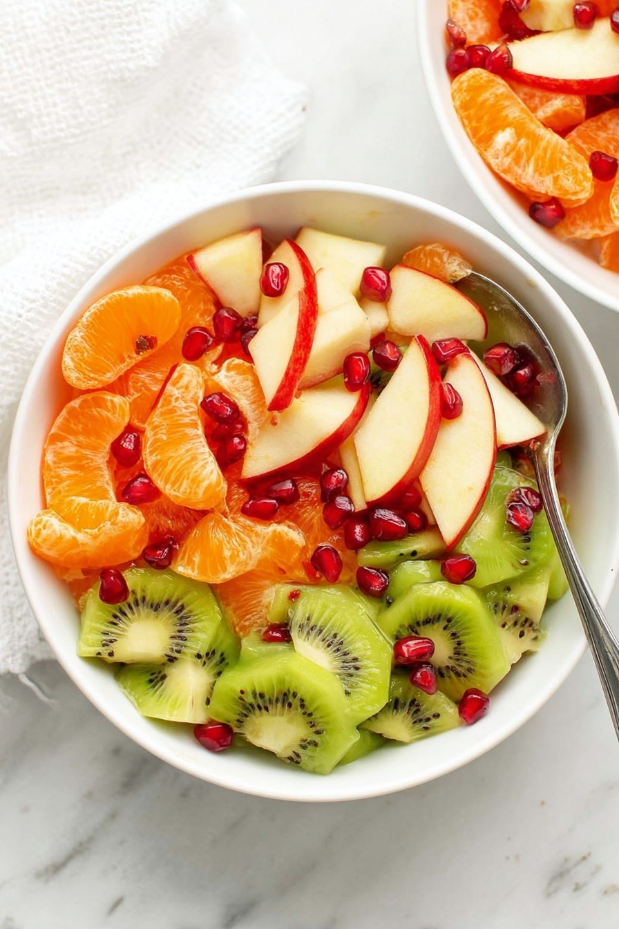A white bowl filled with colorful fruit salad, containing three main layers: at the bottom, slices of green kiwi with black seeds and a smooth texture; in the middle, small wedges of red apple with white flesh and red skin; and on top, bright orange tangerine segments and small shiny red pomegranate seeds scattered evenly. The bowl is placed on a white marbled surface with a silver spoon resting inside. Part of another bowl with similar fruit salad is visible near the top right corner. Photo taken with an iphone --ar 2:3 --v 7