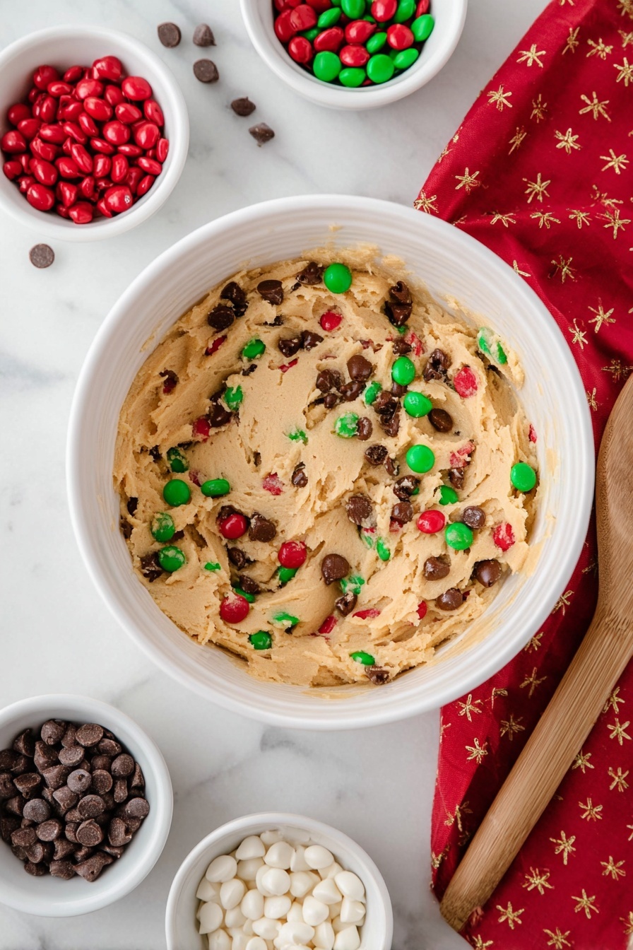 A close-up shows a square cookie with a golden brown color and a slightly cracked surface, held by a woman's hand with black painted nails. The cookie is packed with red, green, and white candy-coated chocolate pieces embedded throughout, alongside small chocolate chips that add texture. The background is softly blurred, with more cookies out of focus, and a white marbled surface beneath. Photo taken with an iphone --ar 2:3 --v 7